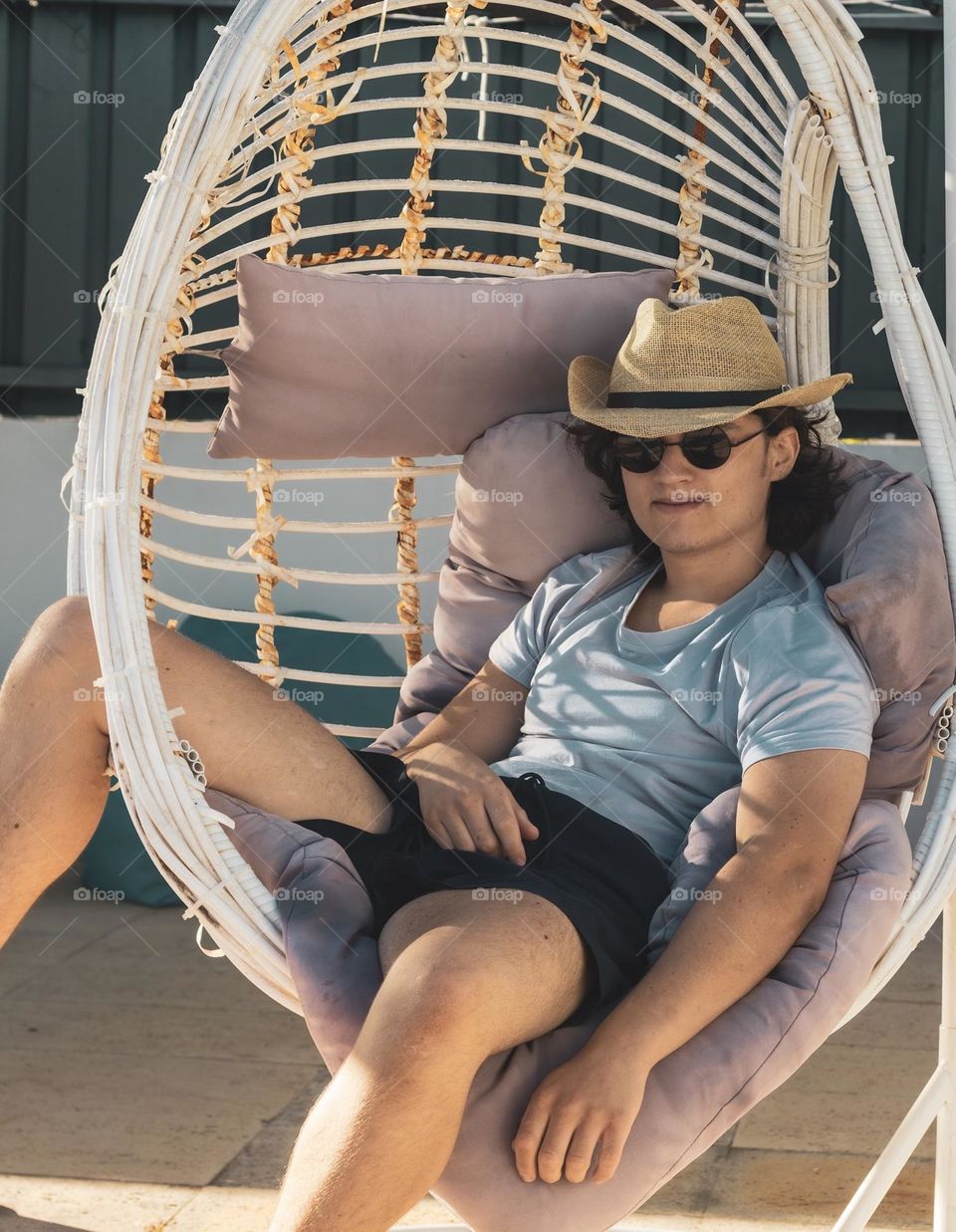 A young man wearing a sun hat & shades sits chilling in a swing chair in the early evening sun