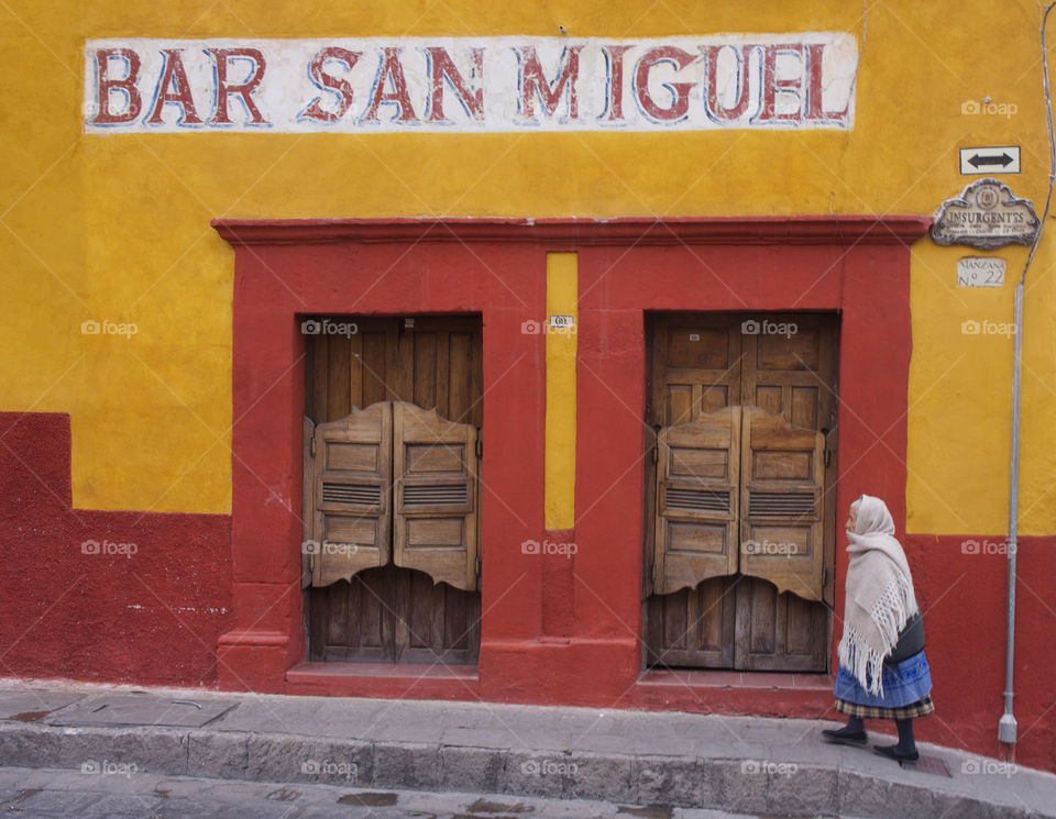 An elderly Mexican  woman is captured walking past the Bar San Miguel in San Miguel de Allende. Mexico.