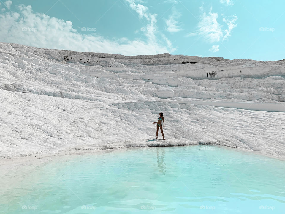 Tiny figure of a young woman in blue bikini standing on white chalk mountain with blue lagoon in front of a blue cloudy sky 