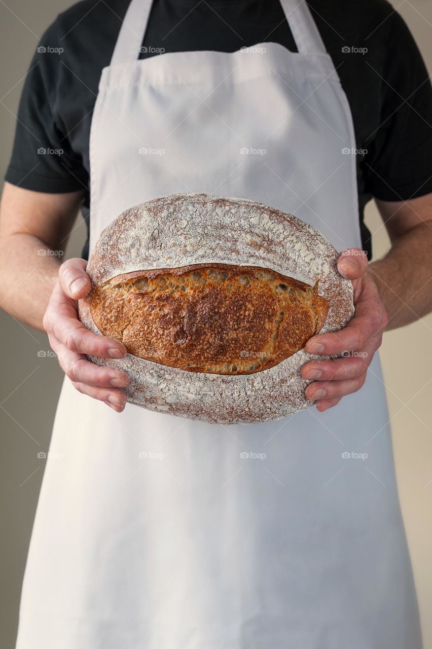 Close-up at bakers hands holding a loaf of sourdough bread in front of him.