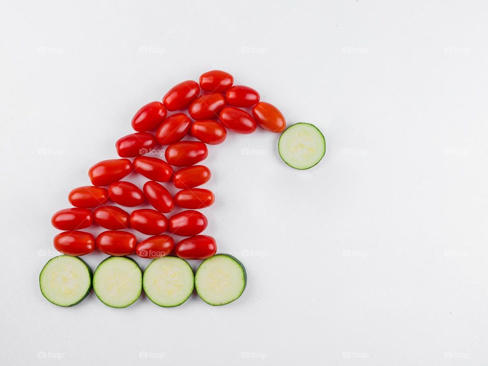 Fresh green zucchini circles and red cherry tomatoes are laid out in the shape of a Santa Claus hat on the left on a white background with copy space on the right, flat lay close-up. Concept vegetables isolates, creative vegetables symbols and signs.