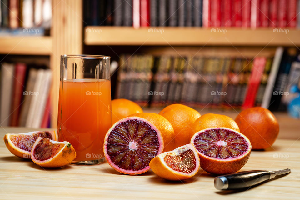 glass of juice, knife and cut red oranges on a light wooden table, blurred bookshelves in the background