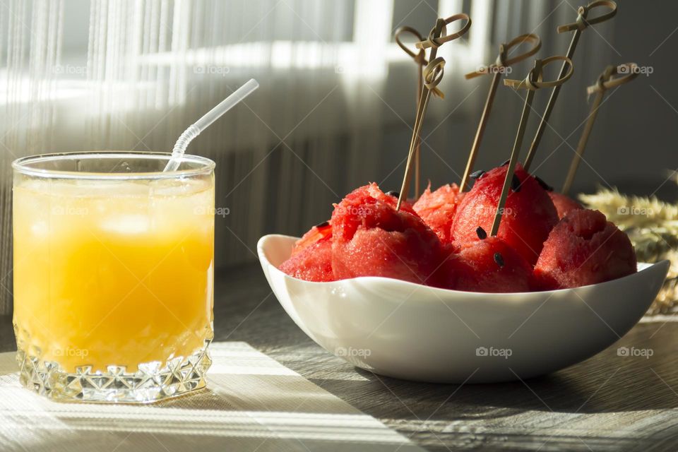 Cooling, orange juice with ice, red watermelon and ears of wheat are on a wooden table in the sun.