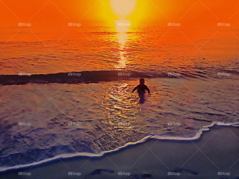 Young boy playing in the ocean enveloped in a sunset ablaze in gold orange and red.