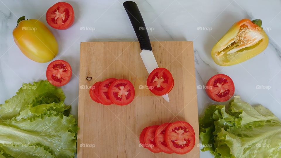 Lunch time! Cutting vegetables for salad. On the kitchen table, a wooden cutting board, a ceramic knife, tomatoes, yellow peppers, lettuce. Cook at home