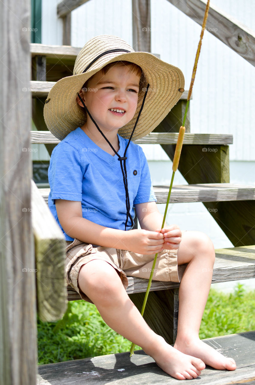 Young boy smiling and sitting on wooden steps outdoors while holding and looking at a cattail