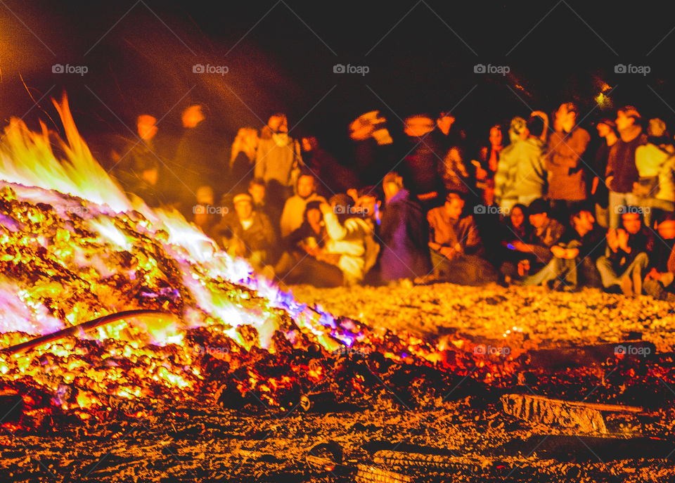 A large group of people gather behind the roaring flames of an autumn bonfire on the beach