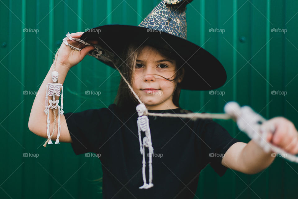 A girl in a witch costume on a green background holds a skeleton in her hands. Halloween concept