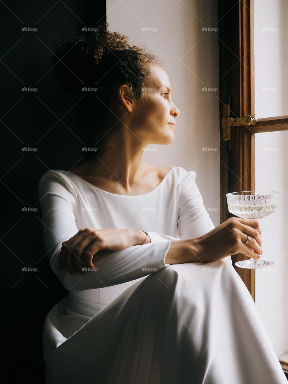 Young beautiful woman with curly hair wearing white dress sits on the window and drinks champagne, portrait of woman, looking at window, lifestyle, at home, relax.