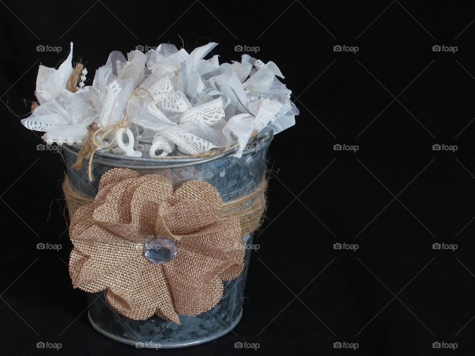 A hand decorated brushed steel bucket with a burlap flower and ribbon filled with hand formed white cloth ribbons against a black background with ample copy space. 