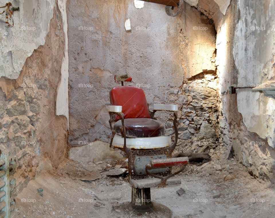 An old red and white rusty barber chair sitting in the middle of a room with crumbled walls and debris all around