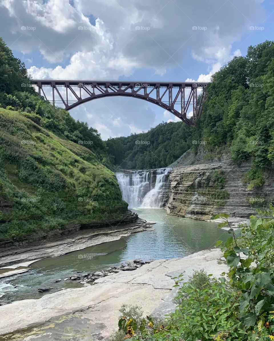 Clear photo of a bridge over a river and waterfall. Has beautiful plants and sky with nice complimenting colors and scenery. 