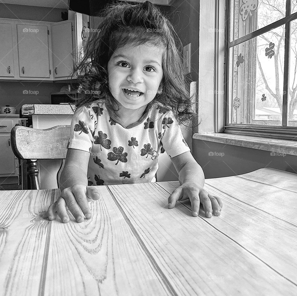 Monochrome portrait of toddler girl, toddler girl waits patiently at the table, black and white portrait of a toddler, happy and smiling toddler girl, dressed for St. Patrick’s Day, smartphone photography, mobile photography, cute toddler smiling 