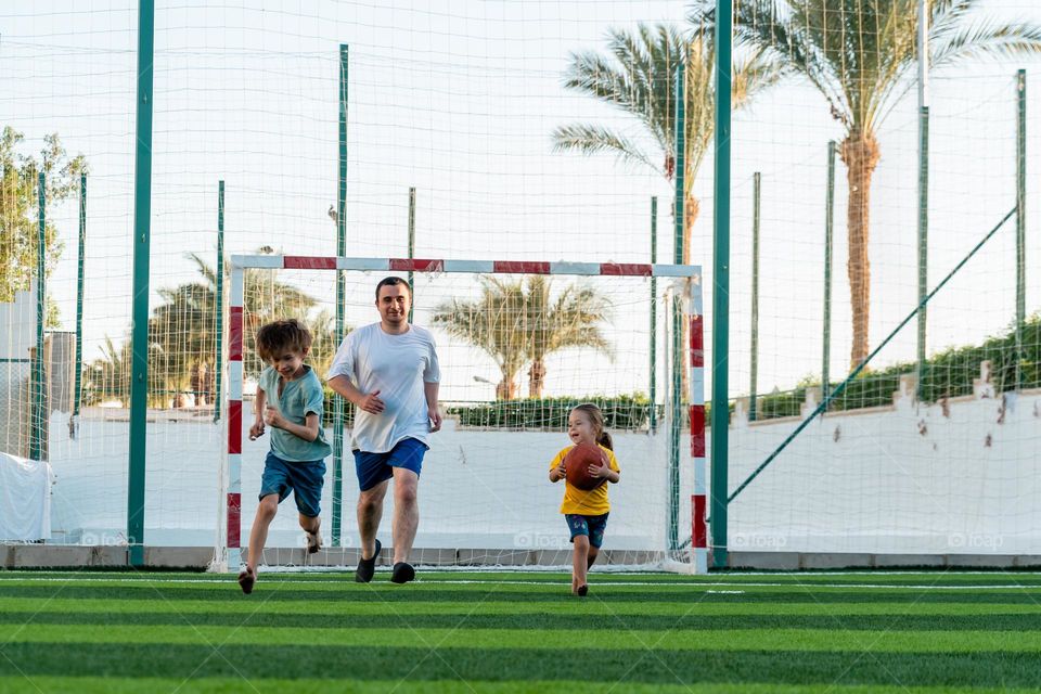 Full body positive man with children running together on sports field to camera during playing football