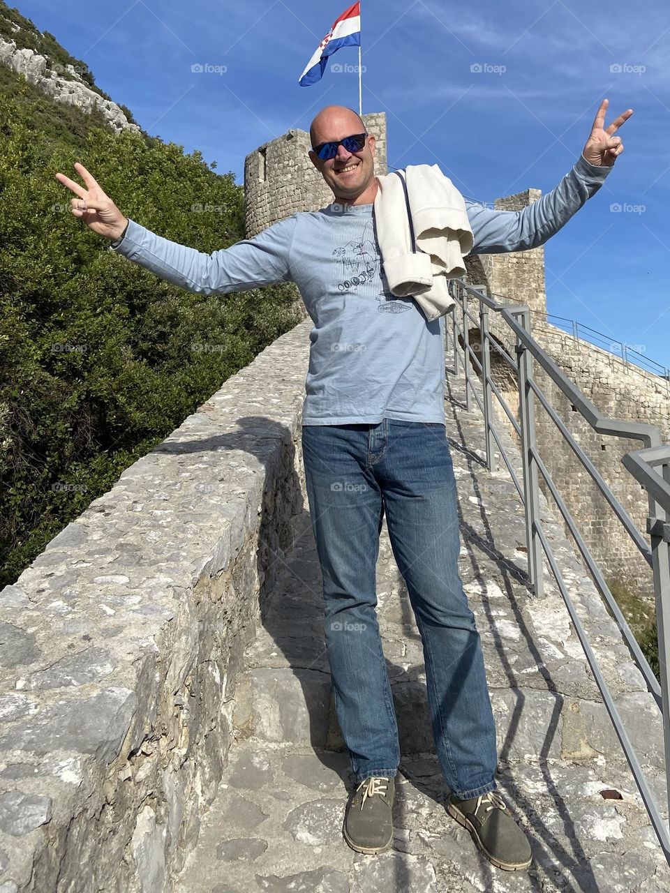 Man celebrating walking up a lot of stairs on the protected walls of Ston, Croatia with a blue sky and Croatian flag in background.