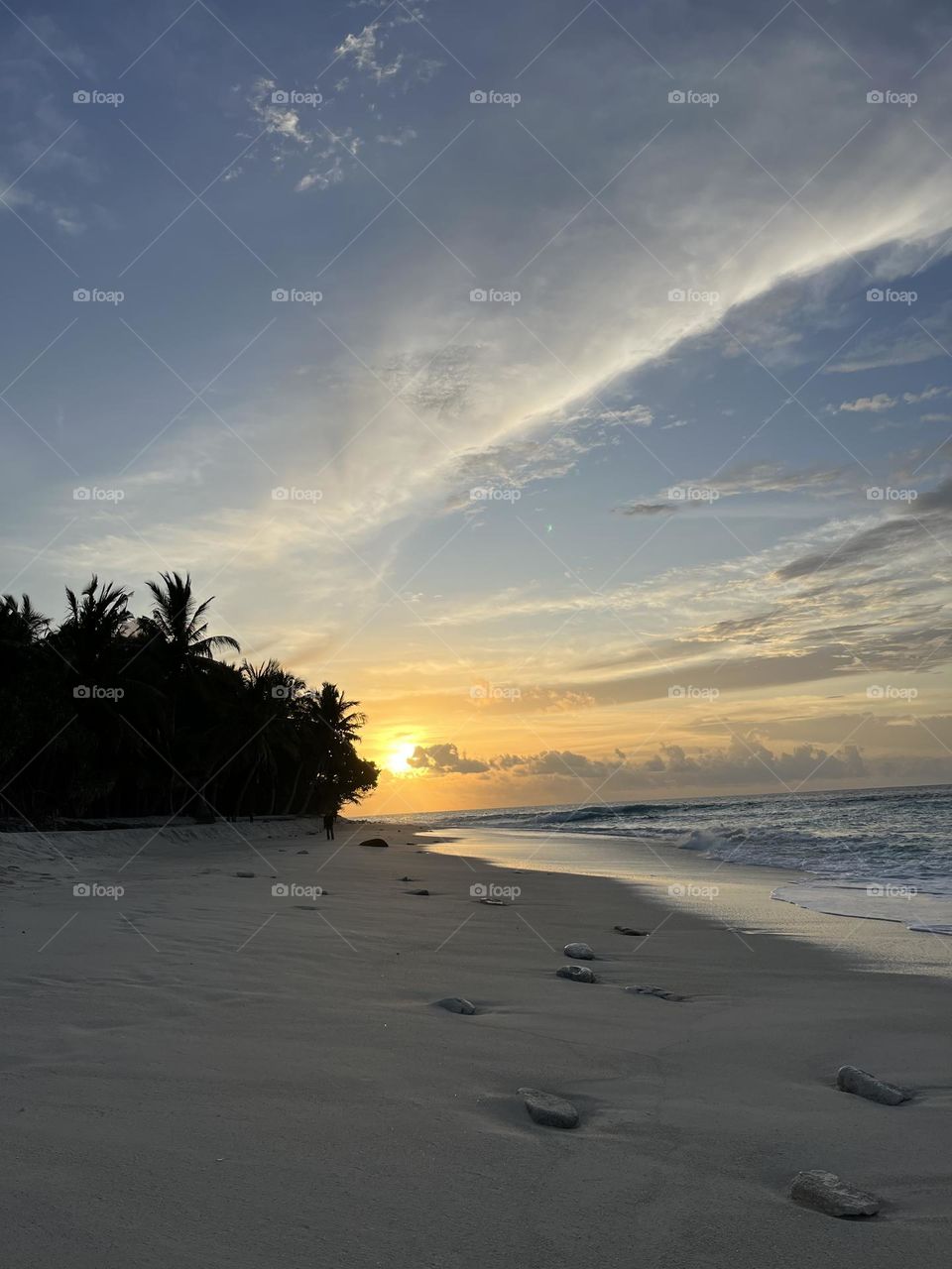 A beautiful sunset on the southern island of Gnaviyani Fuvahmulah in the Maldives. Cotton-like clouds, blue sky and golden rays cover the horizon while mini-waves unhurriedly crawl on the white sandy beach.