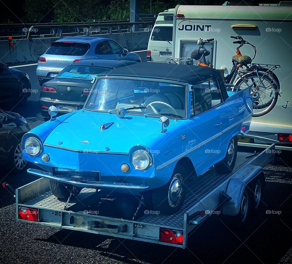 Vacation.  A trailer with a trailer is driving in the stream of cars, on which a blue Amphicar is installed - a West German four-seater floating car.