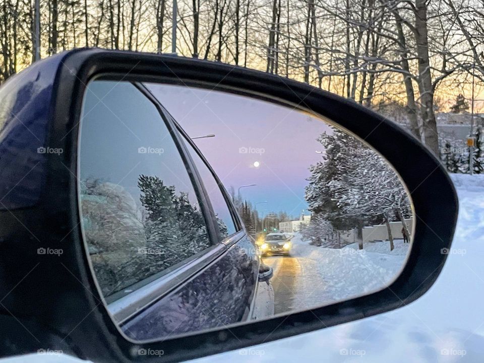 Reflection in the car rearview mirror: cars behind, trees, snow and the full moon on the blue purple lavender sky with the young tree trunks on the orange sunset sky ahead 