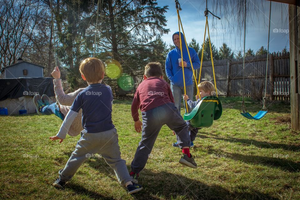 3 boys and their parents play in the backyard on a beautiful spring day, with two of the boys pushing their mother in a swing while dad looks on and drinks.