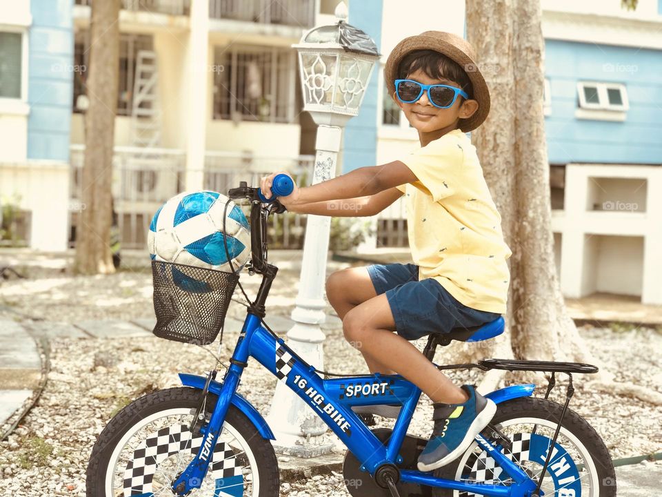 A boy is on bicycle and he has everything blue with him,his by cycle,football, shoes and eye glasses.