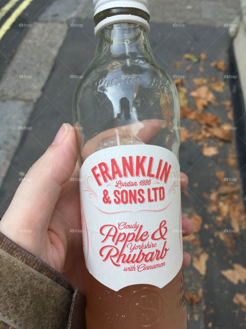 A girls hand holding a Franklin and Sons apple and rhubarb juice in a glass bottle. In the background is a cement street with leaves on the ground 