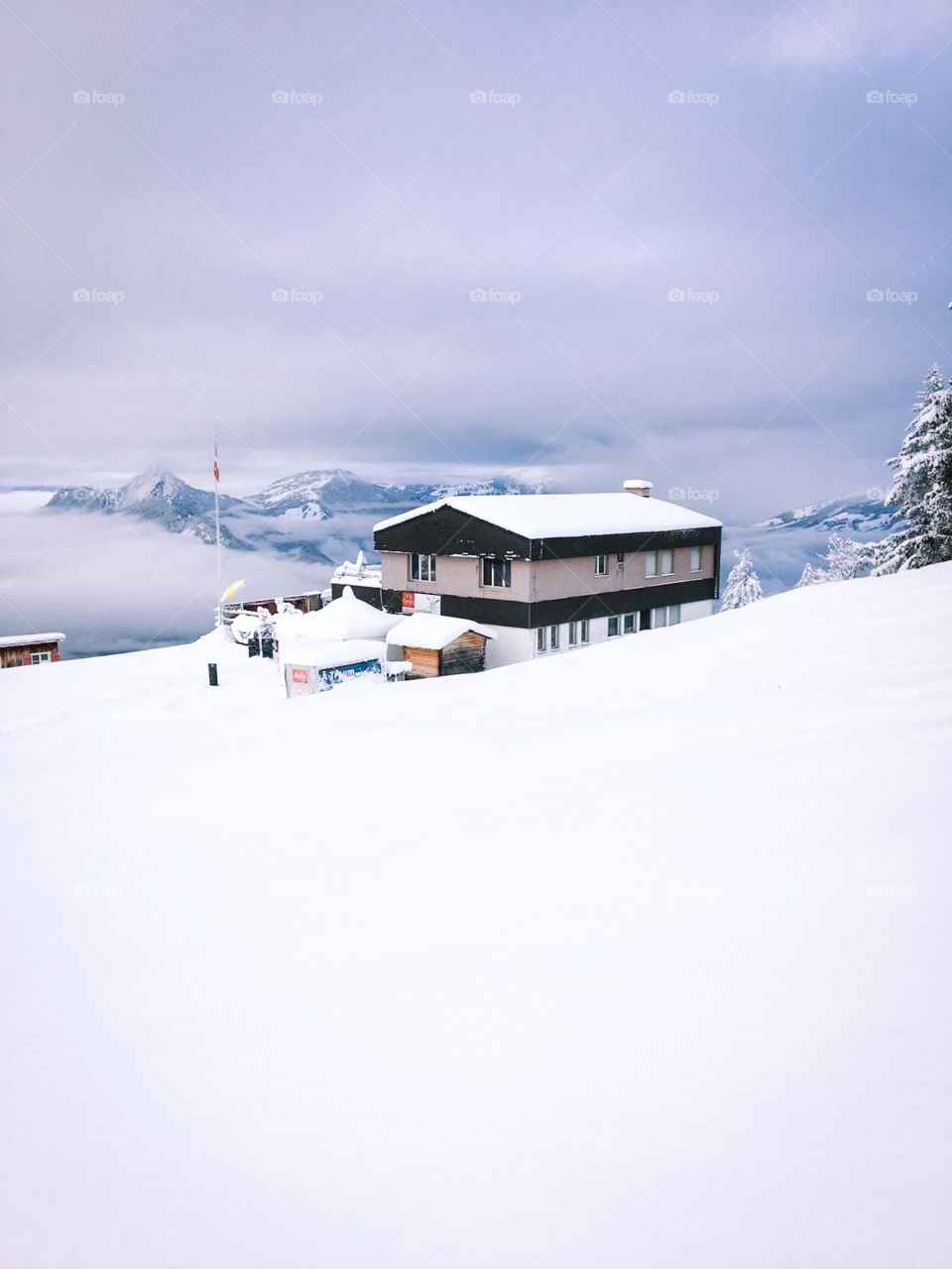 White snow covered the top of the mountain, a house in the middle, around the trees also covered with snow. Seeing a few mountains in the distance, clouds covered the sky, with no sunlight. winter is beautiful but too cold