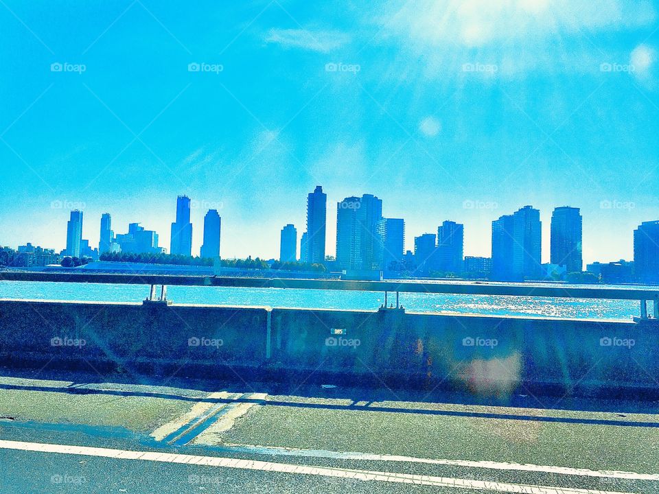 Driving on the highway over one of the bridges connecting Bklyn to LIC, Queens, NY with a stunning view of the Manhattan skyline in the distance. The overhead sun in a perfectly clear blue sky casts refractions in the air. 2020. Hypnotic Productions 