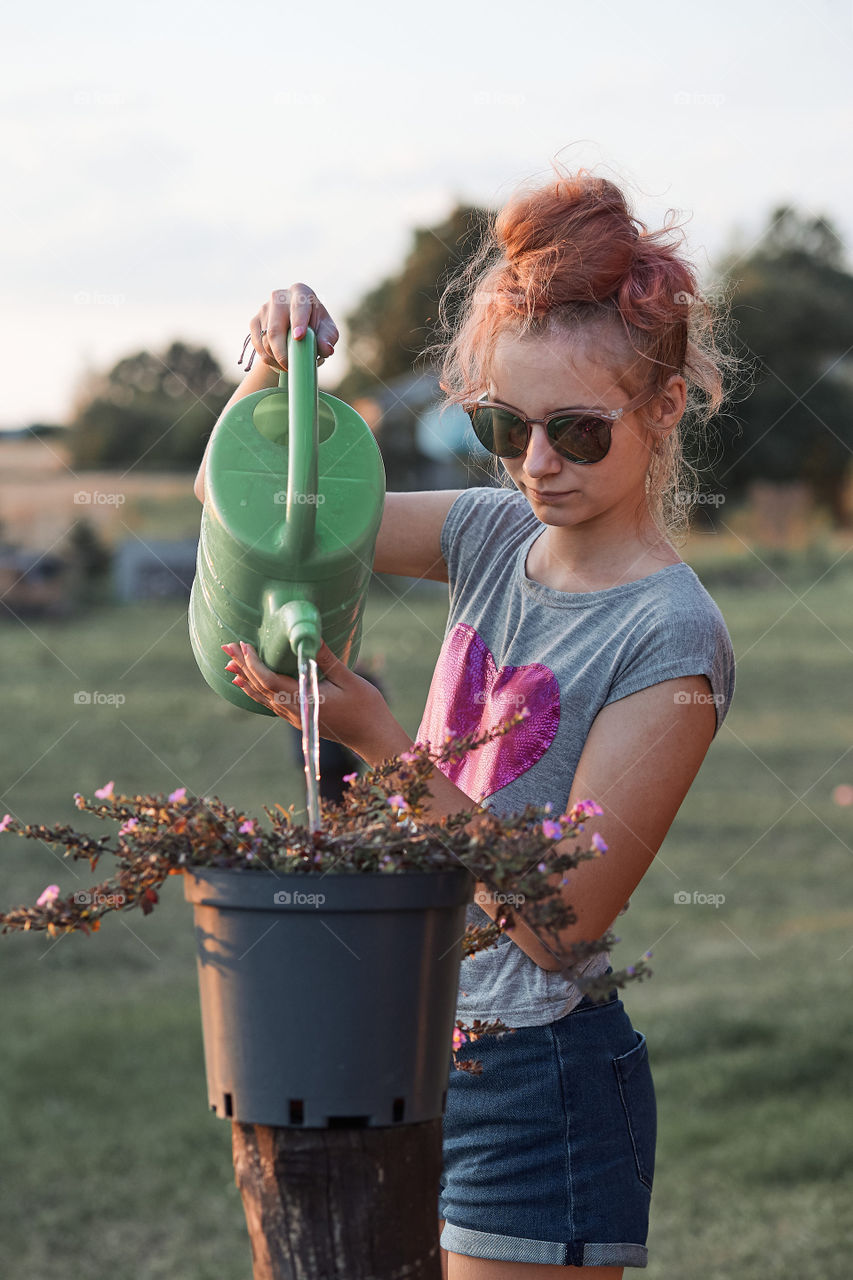 Teenage girl helping to water the flowers growing in flower pot, pouring water from green watering can, working in backyard at sunset. Candid people, real moments, authentic situations