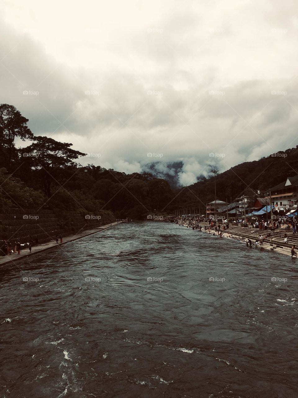 A river flowing calmly through population to deep forest with s cloudy sky over it. 