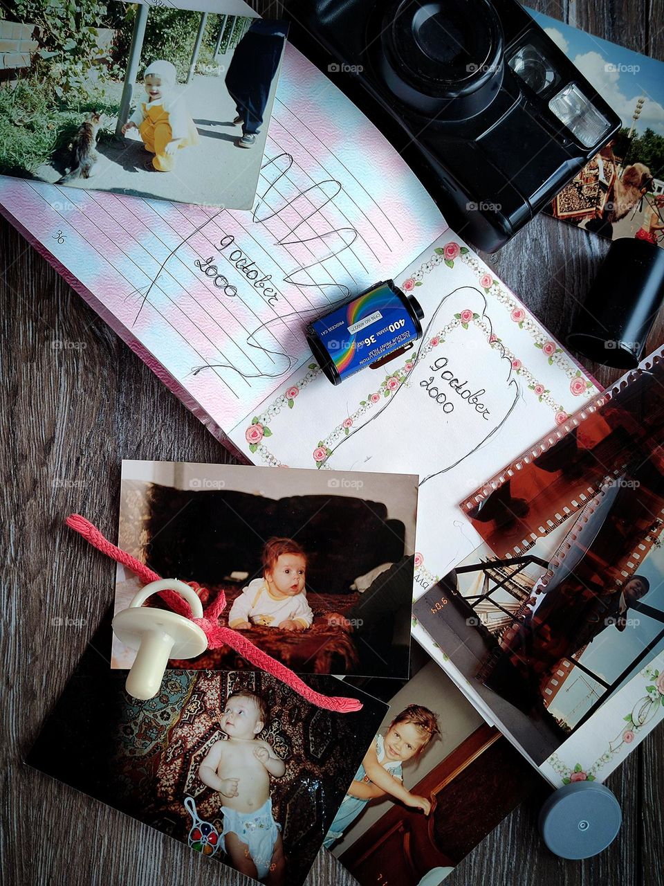On a wooden background lies a notebook with baby foot and hand prints, a pacifier, color photographs of a child, developed films 35 mm, a color film cassette, a camera. Memories