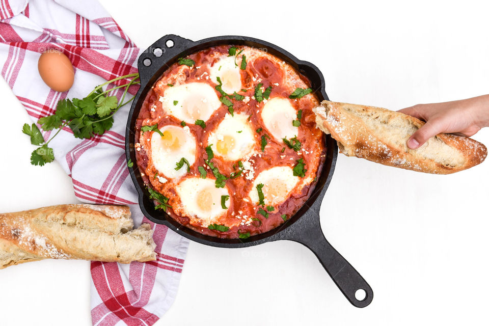 Top view of a shakshouka dish in a cast iron skillet and a person's hand holding a piece of bread