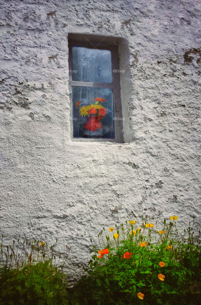 Vase of flowers in a window in an old wall with peeling paint and wild flowers growing on the ground outside against the wall