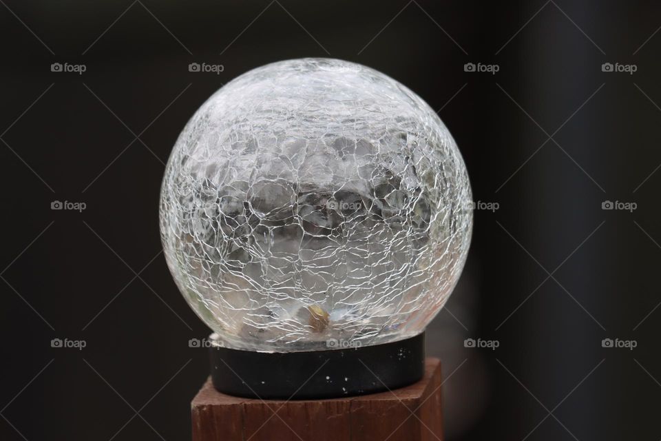 A cracked style glass ball, standing tall on a wooden podium, it’s cracks broadcast to the world and contrasting against the greens of the blurry bush behind it.