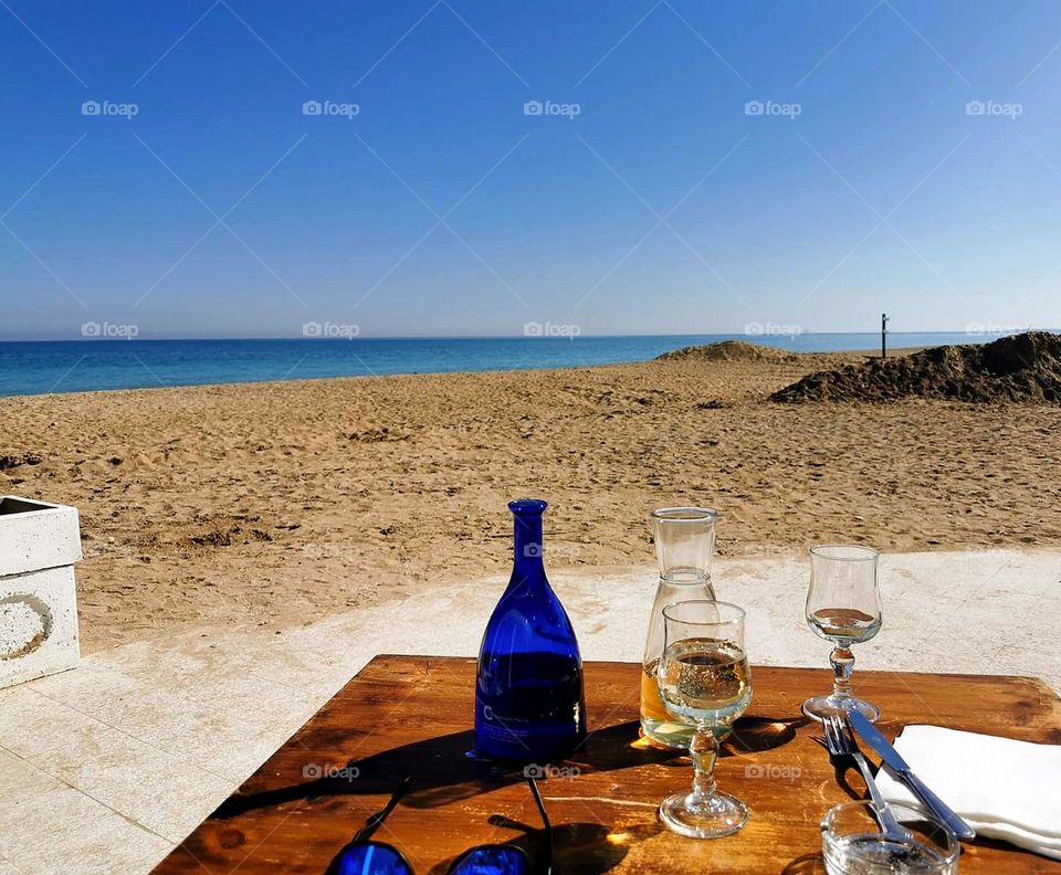 Sea. Sandy shore. The azure sea merges with the horizon. In the foreground is a table with cutlery, two glasses of wine, bottles, sun glasses. Romance
