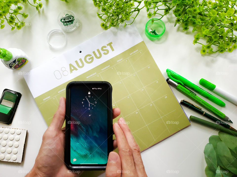 Flatlay:  A man holding his cell phone in front of a green and white calendar for planning, surrounded by green office  stationary and greenery.