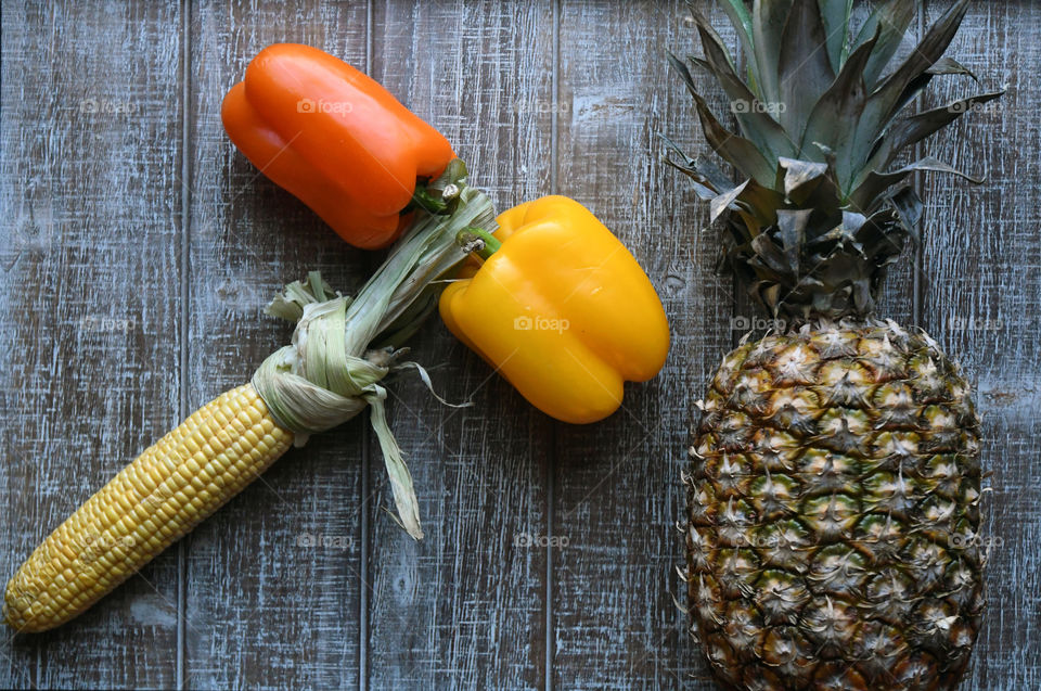 Two bell peppers, orange and yellow with a corn on a cob form to make the shape of a hammer hitting a pineapple.