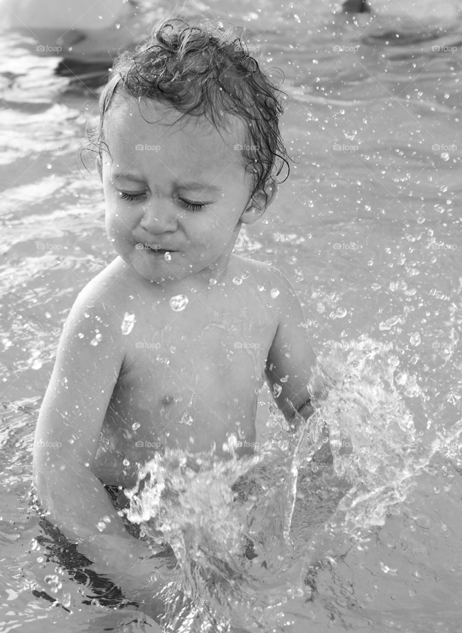 A toddler boy plays and splashes in an outdoor swimming pool on a sunny summer day. 
