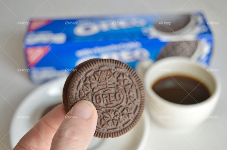 A persons fingers holding an Oreo cookie and a cup of coffee in the background
