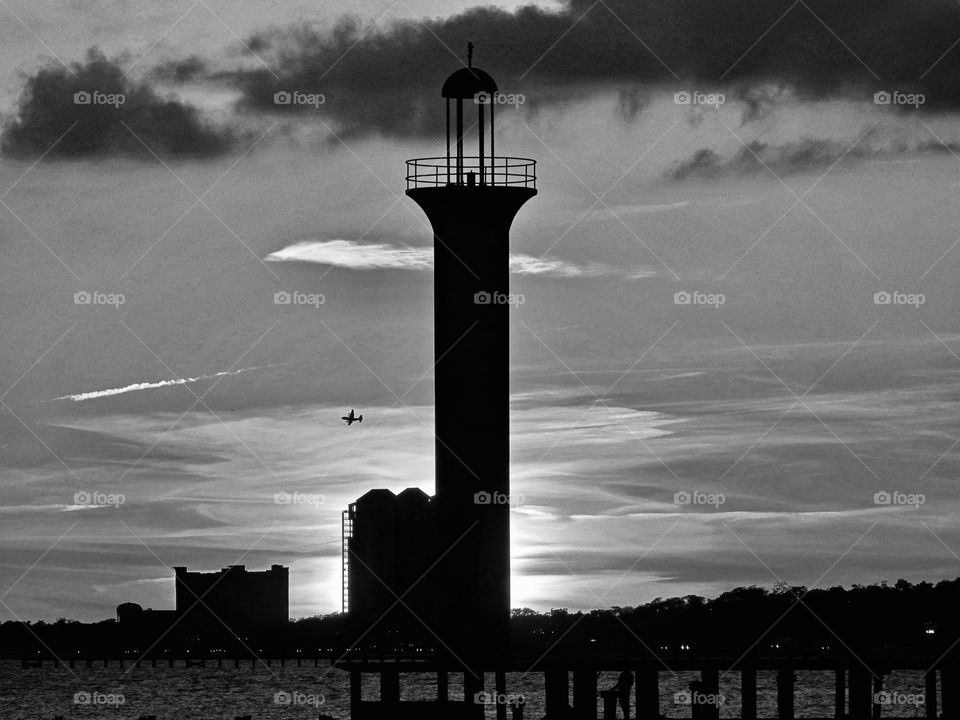 A black and white photo of a lighthouse reaching into the clouds as a plane circles around 