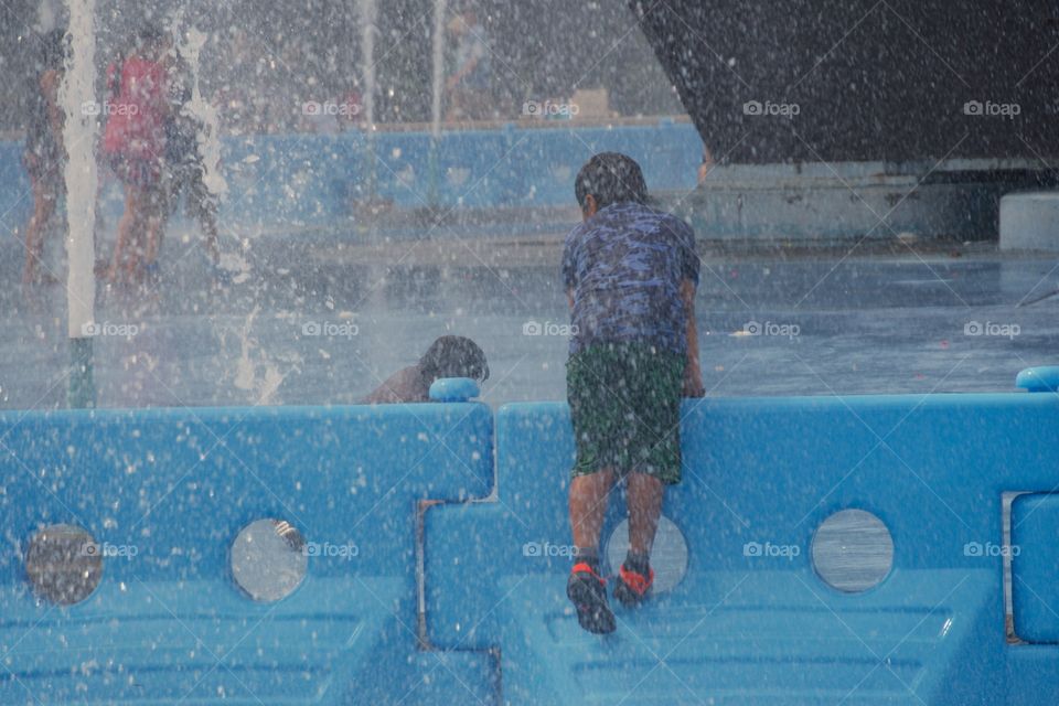 On a hot summer day, children and adults cool off and play in Flushing Meadows Corona Park from the 96  water spray jets  in the fountain which surround the 1964 World's Fair Unisphere landmark, New York City.