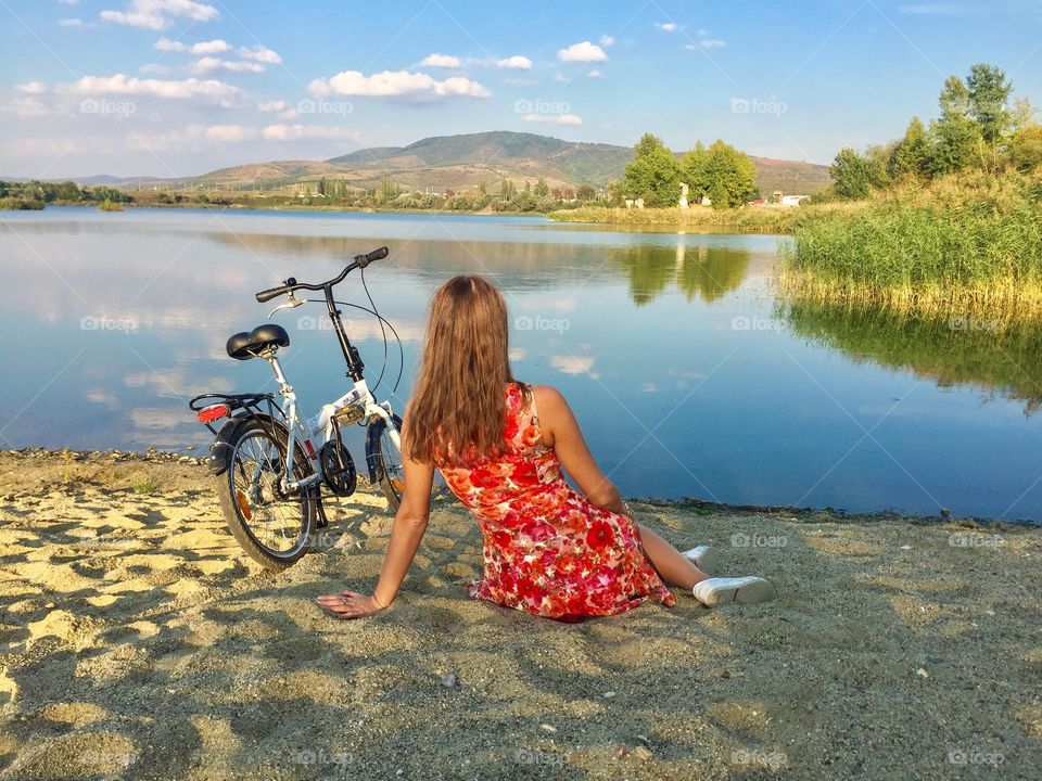 Back of woman wearing flower dress sitting on the sand looking at the lake with bicycle near her