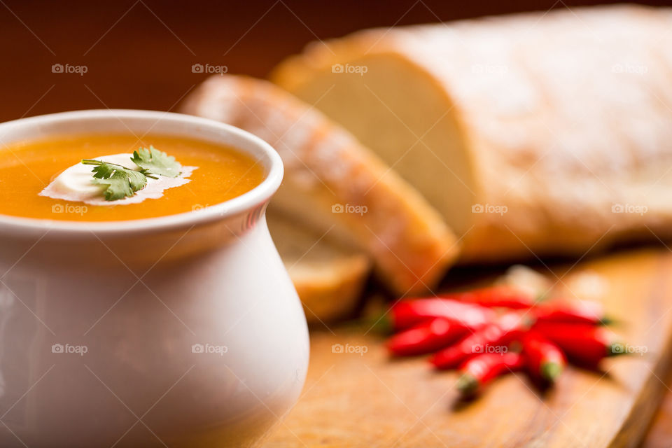 Fresh pumpkin soup with cream and coriander served with fresh bread. Healthy winter soup with added chilli. soup in a white cup on bread board.