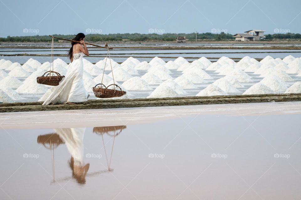 The salt of life. Elegant woman dressed in a long white wedding dress walking along the salt pans with reflection in the salt marshes