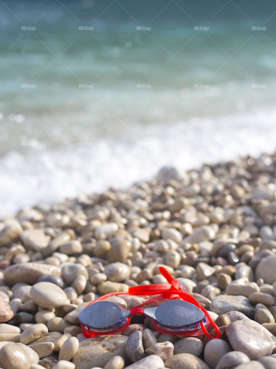 Red swimming goggles lie on the pebble beach near the water