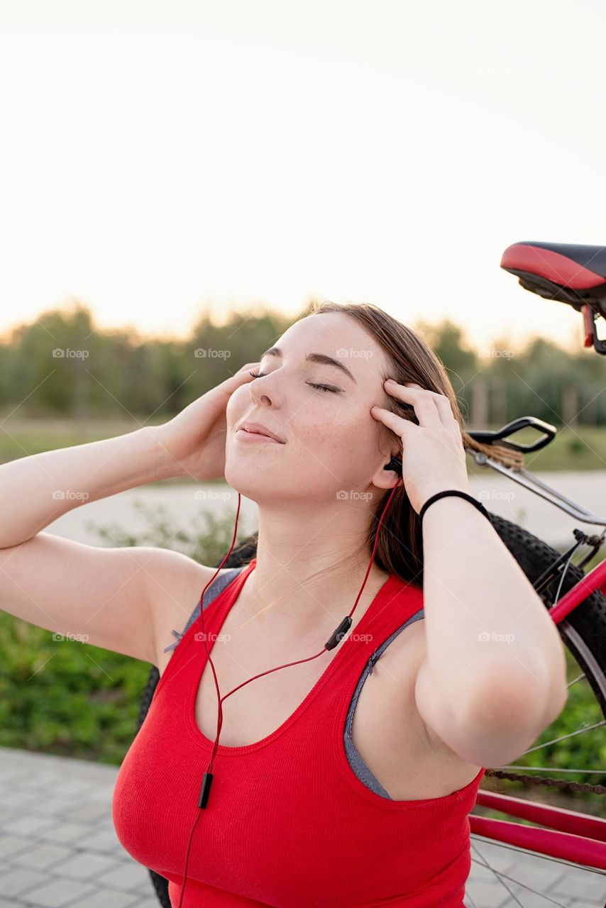 young smiling woman in red shirt and white pants listening to the music standing near her bicycle