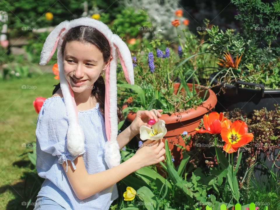 Portrait of a beautiful caucasian teenager girl with a bunny headband on her head sits on a lawn in the garden, looking away with a smile on her face, hides a chocolate easter egg in a shiny wrapper in a white tulip growing near pots of flowers on a