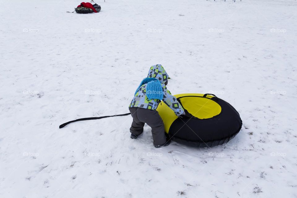 A small, carefree boy walks in the white snow in winter and rides a tubing in the park, near trees in the snow.