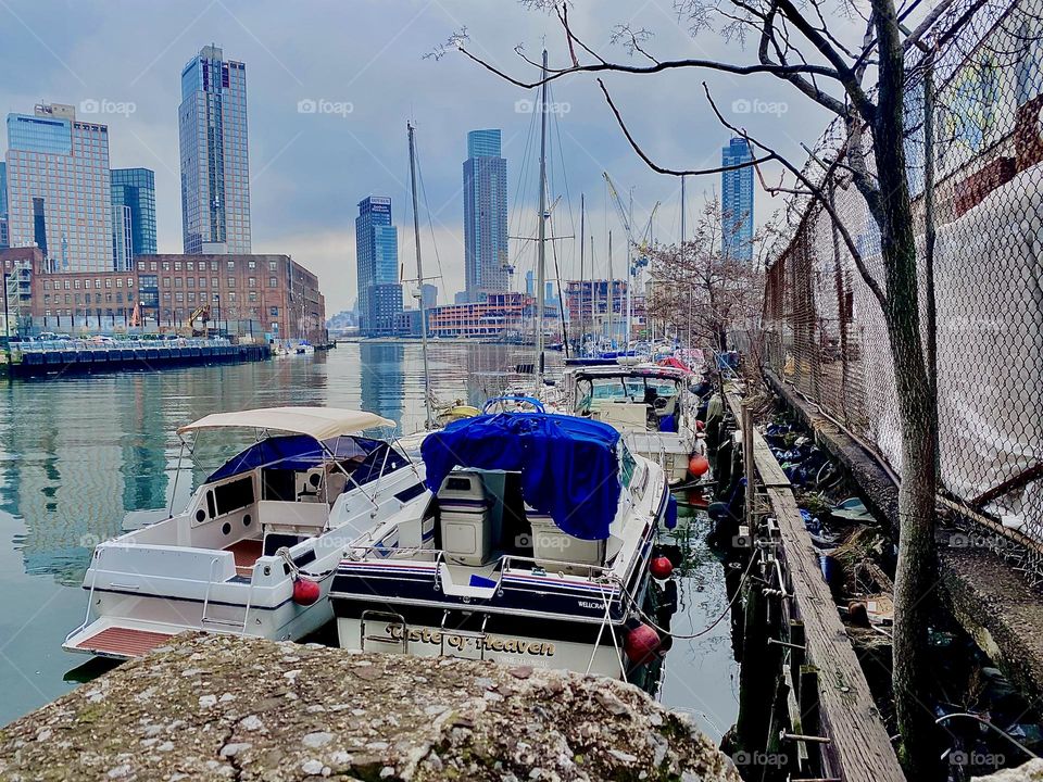 The boats at “Newtown Creek” closeup, a tree lined wall on the right on the LIC shore, “Greenpoint”, Bklyn across the water on the left and the “Manhattan” skyline in the far distance, this is an all around great view. 2023. Hypnotic Productions