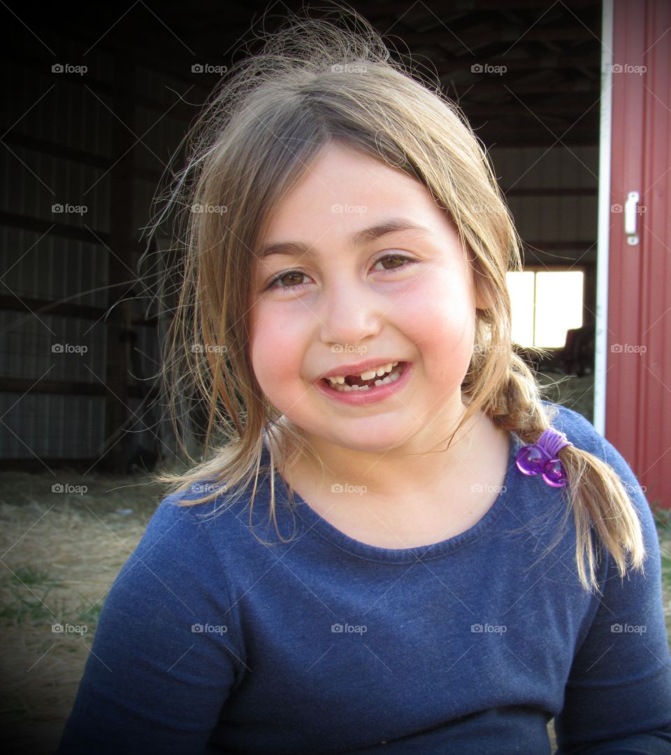 Portrait of a six year old girl smiling and missing a tooth sitting outside of a red barn in the spring