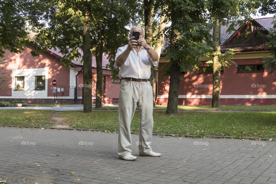 An elderly man walks alone in the park in the summer. A modern pensioner, businessman in a white shirt and trousers takes pictures with a camera in a mobile phone.